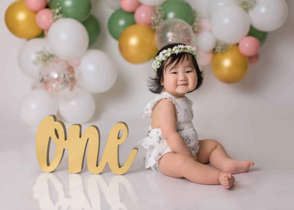 A one-year-old baby wearing a floral romper and a flower crown, sitting next to a golden "One" sign. The backdrop features pastel green, pink, white, and gold balloons.