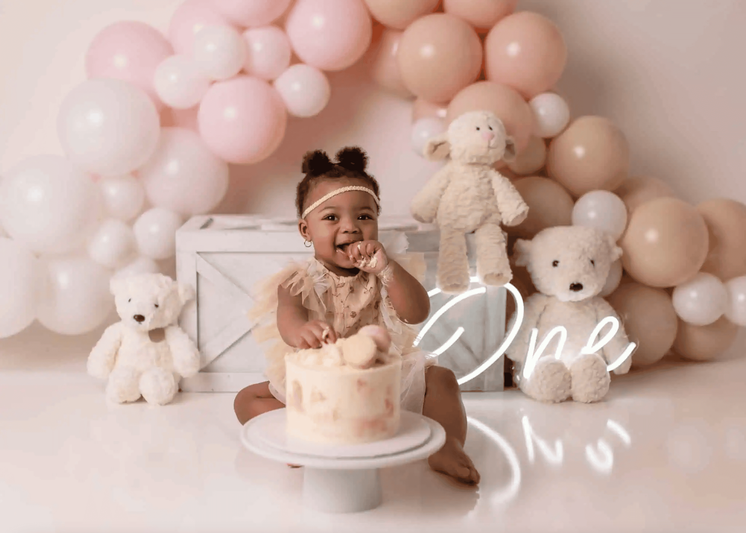 Adorable baby celebrating their first birthday with a cake smash session in Edmonton. The baby is dressed in a cute beige outfit, surrounded by pastel pink and beige balloon decorations, plush teddy bears, and a delicious birthday cake