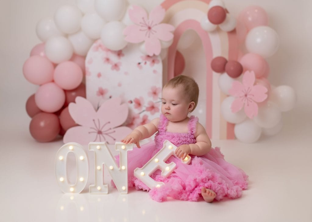 Edmonton cake smash photo featuring pink flowers, white ballons, and a dainty setup for a baby girl’s first birthday.