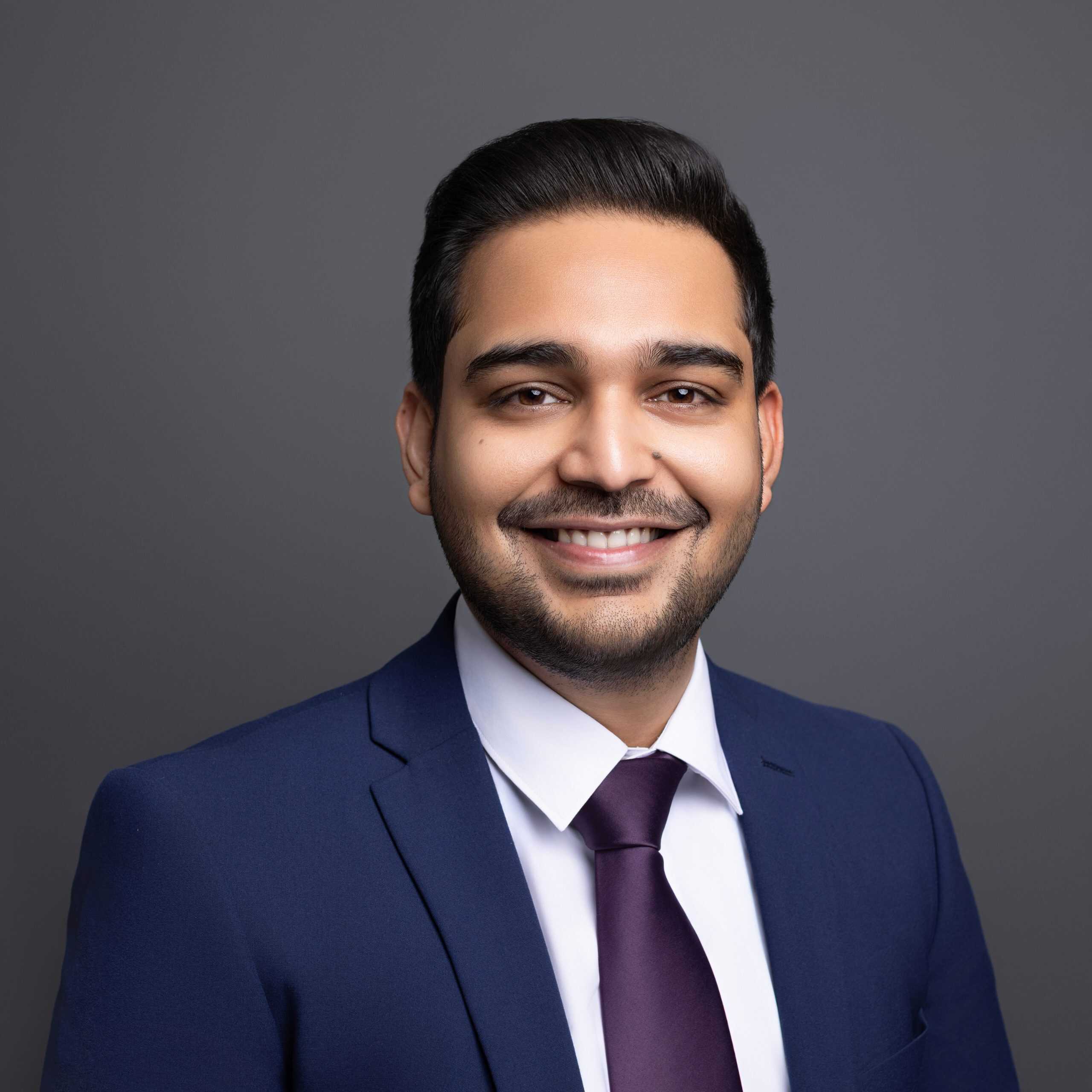 Professional business headshot of a confident man wearing a navy blazer and white blouse, smiling against a neutral gray background in Edmonton.